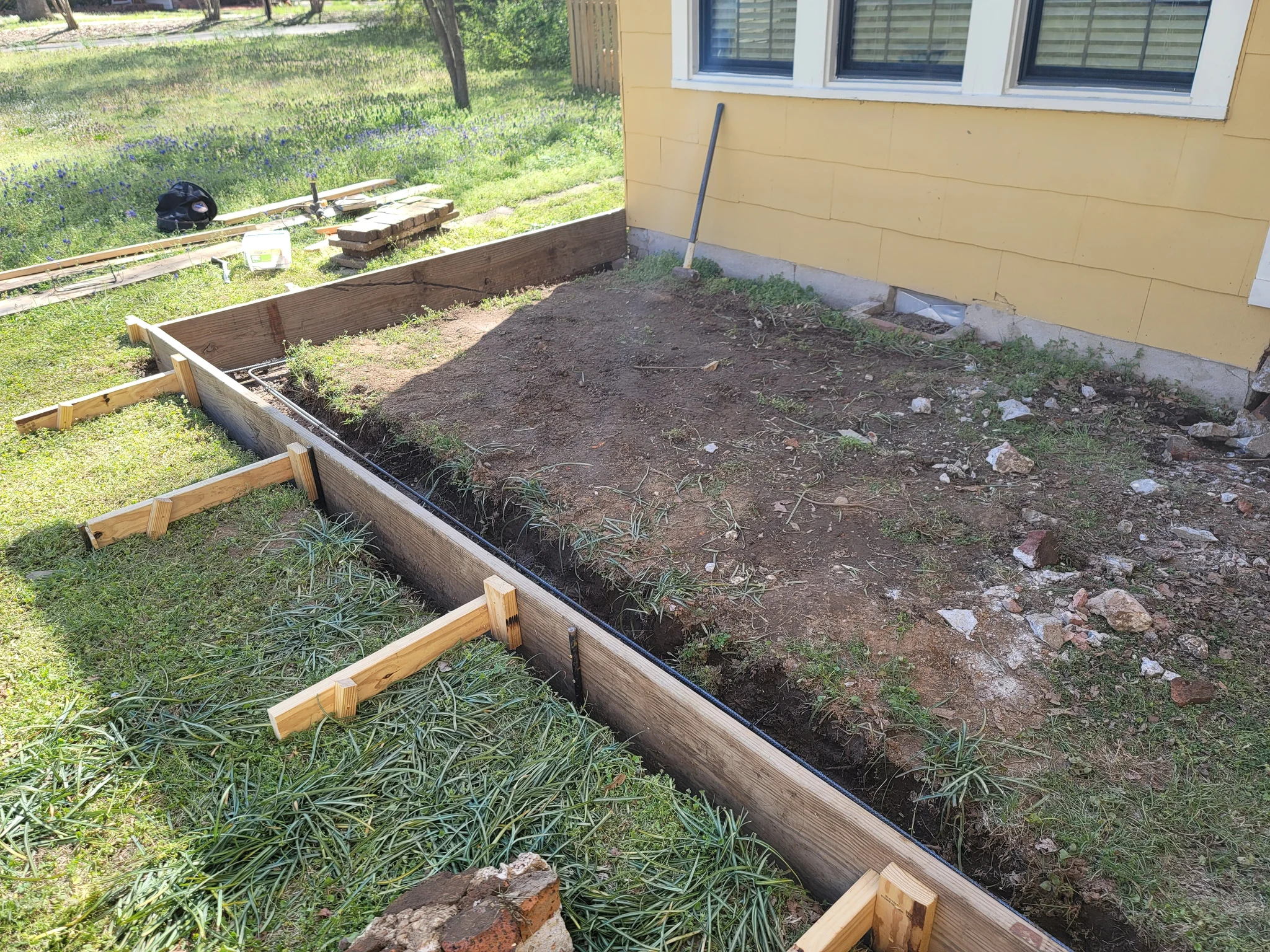 Excavated ground with formwork staked out for a new slab beside a yellow house.