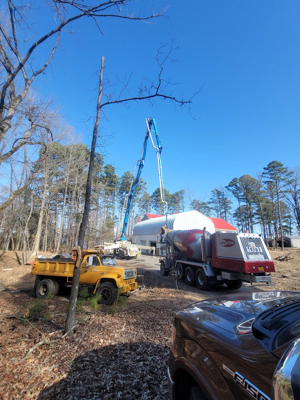 A concrete pump truck and a dump truck staged among the trees on delivery day.