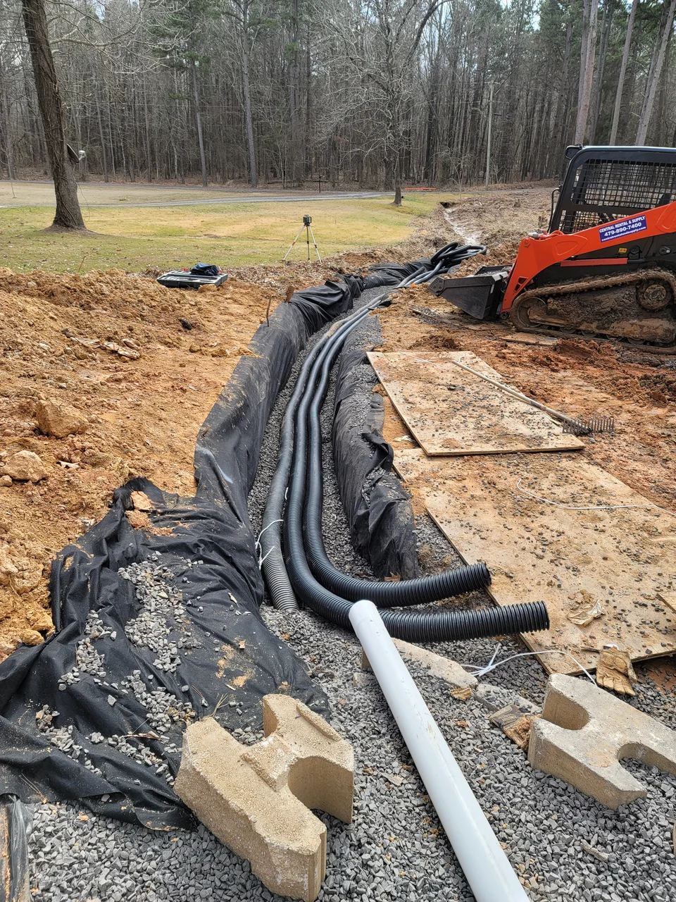 Bundled conduit and pipe laid over fabric in a gravel bed, with a skid steer working behind.