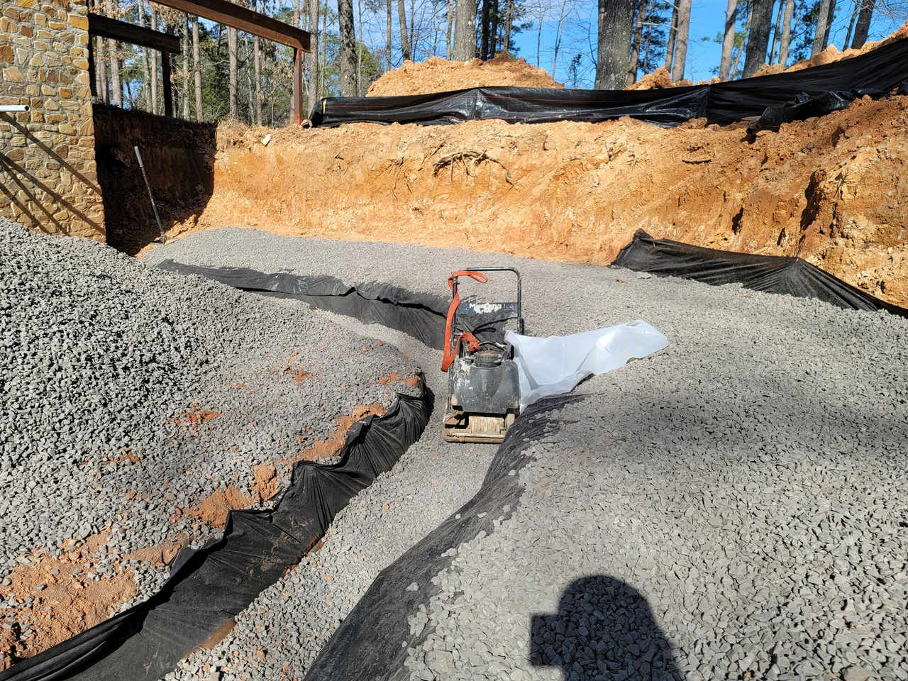 A plate compactor working gravel over a fabric-lined drainage run.